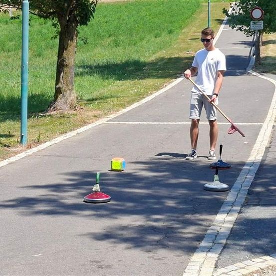 Ein Mann in einem weißen T-Shirt und Sonnenbrille spielt auf einem gepflasterten Weg mit einem Stock und drei Objekten ein Spiel.