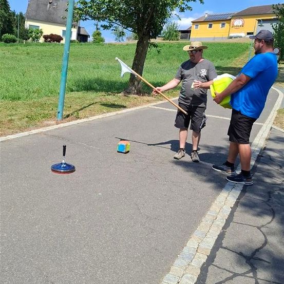 Zwei Männer stehen auf einer Straße, einer hält einen Stock mit einer Flagge und der andere einen gelben Eimer. Ein kleiner Würfel liegt auf der Straße.