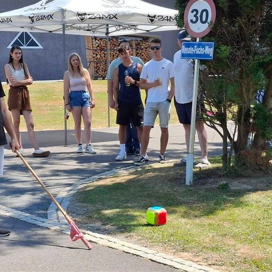 Eine Gruppe von Menschen steht auf einer Straße, ein Mann hält einen Stock und scheint ein Spiel zu spielen. Ein Geschwindigkeitslimit-Schild und ein Straßenschild befinden sich auf der rechten Seite.