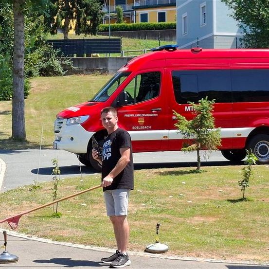 Ein Mann in einem schwarzen T-Shirt steht auf einem Bürgersteig, hält einen Stock und einen Pokal in der Hand, mit einem roten Feuerwehrauto im Hintergrund.