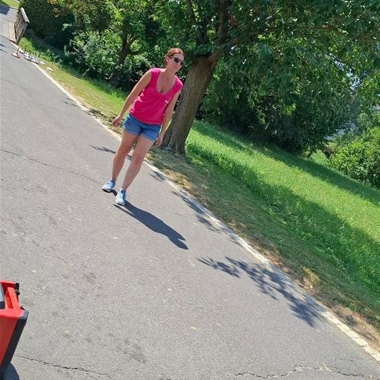 Eine Frau in einem rosa Oberteil und Jeansshorts skateboardet auf einer asphaltierten Straße mit einem Baum und einem Grasbereich an der Seite.