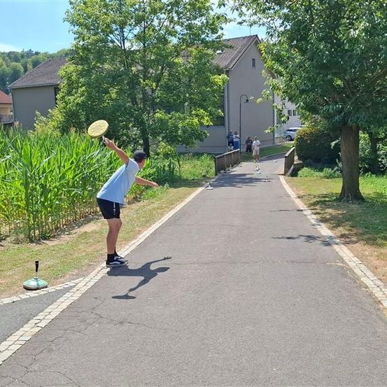Ein Mann wirft einen Frisbee auf einem von Grün und Häusern umgebenen Pfad.