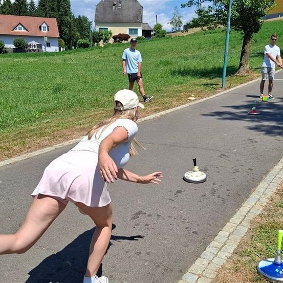 Eine Frau in einem rosa Kleid spielt Cornhole auf einem gepflasterten Weg. Zwei Männer beobachten sie.
