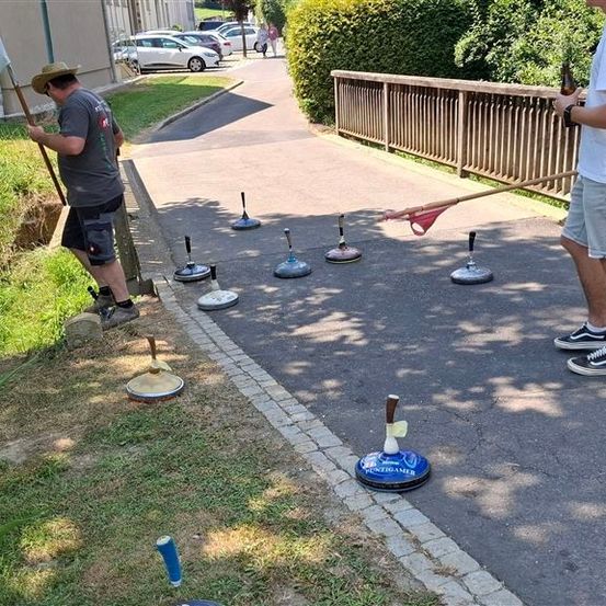 Zwei Männer spielen ein Kegelspiel auf einem gepflasterten Weg, mit mehreren Kegelpins verstreut. Ein Mann hält eine Flasche und einen Kegelschläger, während der andere Mann im Begriff ist, seinen Kegelschläger zu werfen.