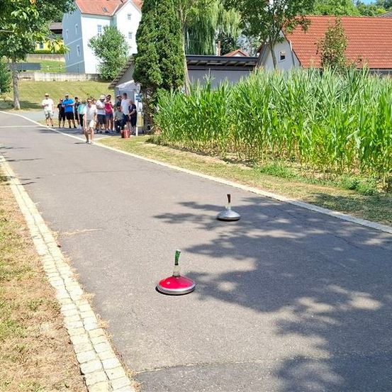 Eine Gruppe von Menschen steht auf einer Straße mit zwei roten und silbernen Objekten darauf. Sie sind von hohem Gras und einem Maisfeld umgeben. In der Ferne befinden sich Häuser und Bäume.