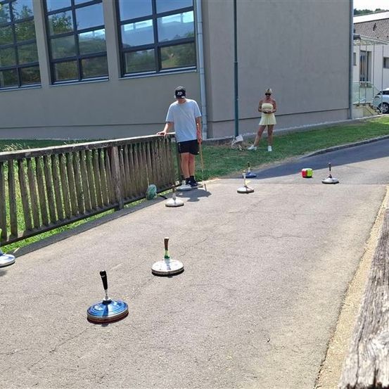 Zwei Personen spielen ein Spiel mit Garten-Dart auf einer gepflasterten Fläche, wobei eine Person sich darauf vorbereitet, zu werfen, während die andere in der Nähe steht. Die Umgebung umfasst einen Holzzaun, ein Gebäude und ein geparktes Auto.