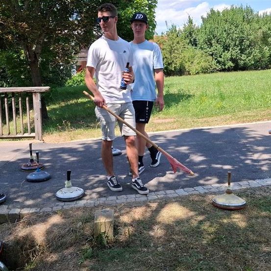 Zwei Männer gehen mit einem Besen und einer Flasche auf einem Pfad, umgeben von verschiedenen Gegenständen.