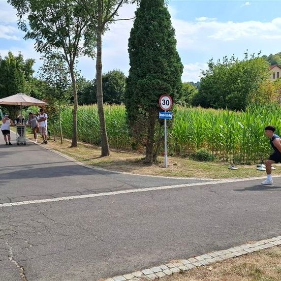 Ein Mann joggt auf einer Straße mit einem Geschwindigkeitsbegrenzungsschild von 30 km/h. Eine Gruppe von Menschen steht in der Nähe eines Zeltes und eines Tisches mit einer Flasche darauf.