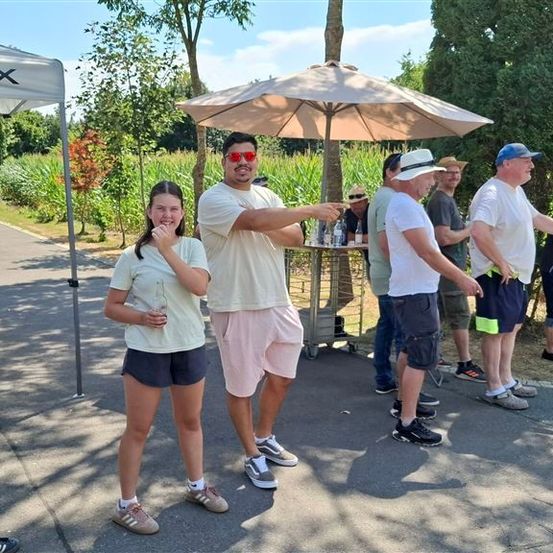 Eine Gruppe von Menschen steht unter einem Regenschirm, einer Mann zeigt und eine Frau lächelt. Sie sind in einem Park mit einem Geschwindigkeitsbegrenzungsschild und einem Zelt.
