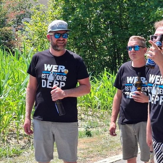 Vier Männer in gleichen T-Shirts stehen auf einem Feld, jeder hält eine Flasche Bier. Sie tragen Sonnenbrillen und Freizeitkleidung.