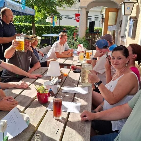 Eine Gruppe von Menschen sitzt um einen Holztisch im Freien, einige halten Gläser mit Bier und andere führen Gespräche. Die Umgebung scheint ein Garten oder ein Sitzbereich im Freien zu sein.