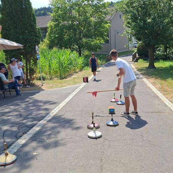 Eine Gruppe von Menschen spielt ein Spiel auf einem gepflasterten Weg mit einer roten Fahne und einem Stock. Ein Mann steht und hält einen Stock, während ein anderer Mann auf einem Stuhl sitzt.