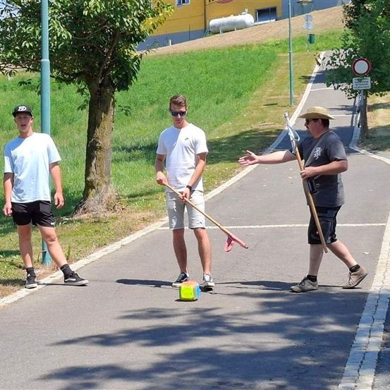 Drei Männer stehen auf einem Pfad, halten Paddel und spielen ein Spiel. Der Mann auf der linken Seite trägt eine Mütze und Turnschuhe, während der Mann in der Mitte eine Sonnenbrille und Turnschuhe trägt. Der Mann auf der rechten Seite trägt eine Mütze und hält ein Paddel.