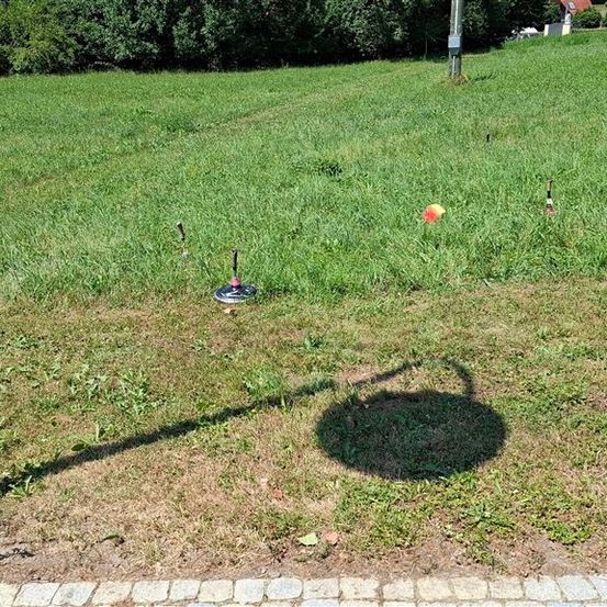 Eine Person steht auf einem Grasfeld mit mehreren aufgestellten Wurfzielen. Die Ziele sind aus Metall und haben eine kreisförmige Form. Die Person trägt Turnschuhe und hält ein Wurfobjekt. Der Schatten der Person wird auf den Boden geworfen.