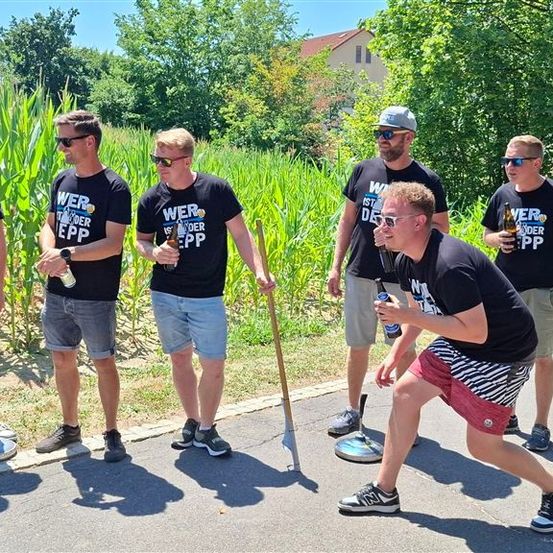 Eine Gruppe von Männern in identischen T-Shirts steht in einem Maisfeld und hält Flaschen und Stöcke. Sie scheinen sich auf ein Spiel vorzubereiten.