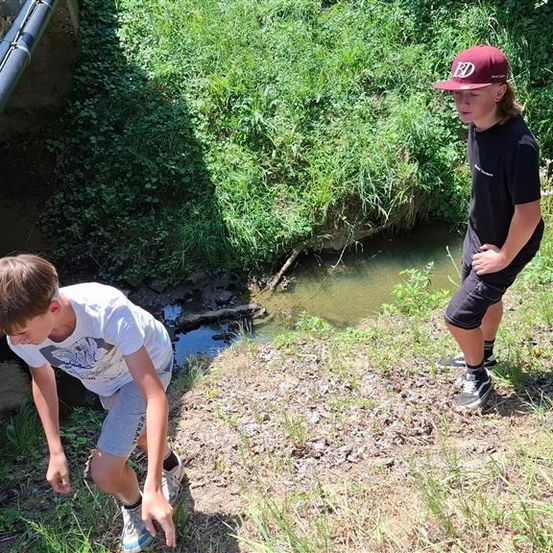 Zwei Jungen stehen am Rand eines kleinen Baches. Ein Junge schaut ins Wasser, während der andere Junge mit den Händen auf den Hüften steht.