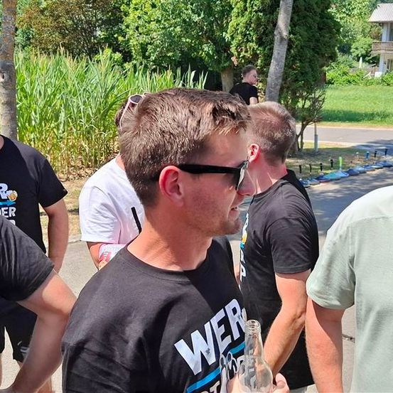 Eine Gruppe von Männern in schwarzen und weißen T-Shirts steht draußen, einer hält eine Flasche. Sie lächeln und scheinen ein Gespräch zu führen. Dahinter befindet sich ein Rasen mit Bäumen und einem Haus.