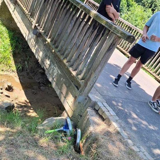 Zwei Personen stehen auf einer Holzbrücke, eine hält sich an der Brüstung fest, während die andere etwas auf dem Boden betrachtet. Einige zerbrochene Gegenstände liegen auf dem Boden.
