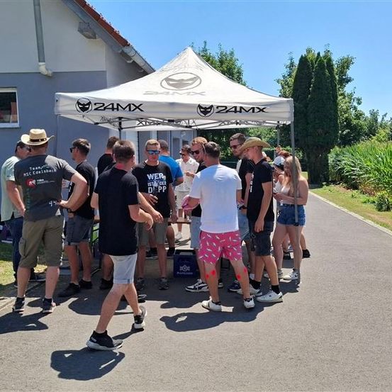 Eine Gruppe von Menschen versammelt sich unter einem weißen Baldachin mit dem 24MX-Logo, einige tragen Sonnenbrillen und Turnschuhe, vor einem Gebäude mit einem Fenster und einem roten Auto im Inneren.