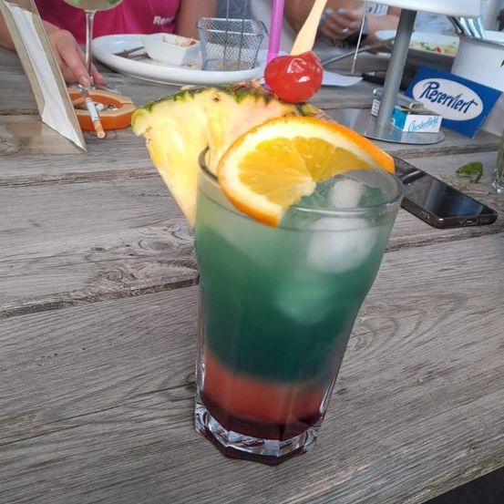 Ein farbenfroher Cocktail mit einer Ananasscheibe und einer Orangenscheibe obenauf, auf einem Holztisch.