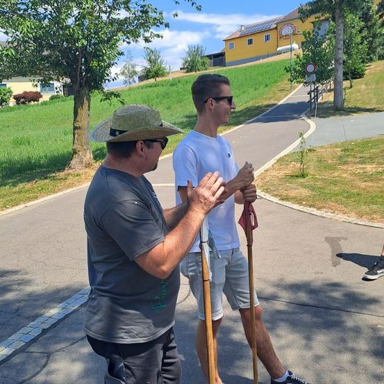 Zwei Männer stehen auf einem Pfad, einer trägt einen Hut und hält einen Stock, der andere ein weißes T-Shirt und Shorts, beide lächeln. Hinter ihnen befindet sich ein grasbewachsener Hügel mit einem gelben Gebäude.