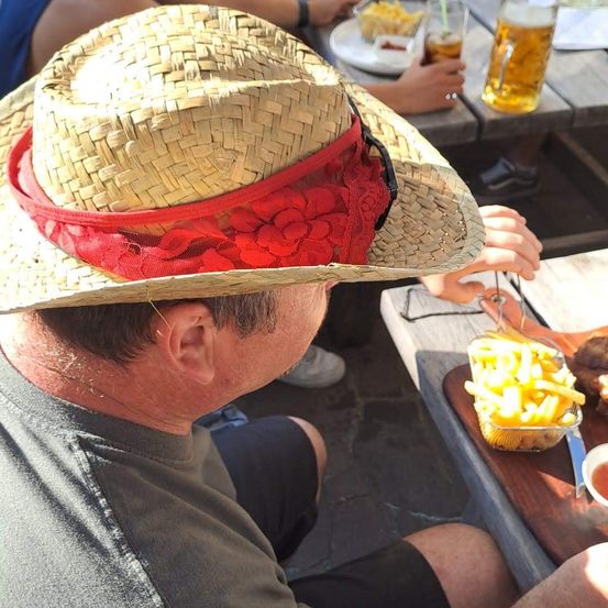 Ein Mann mit einem Strohhut mit rotem Band und Spitze sitzt an einem Tisch mit einem Teller Pommes und einem Bier.