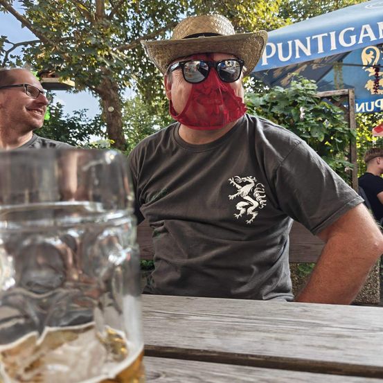 Ein Mann mit einer roten Gesichtsmaske und einem Strohhut sitzt an einem Tisch mit einem Glas Bier. Ein weiterer Mann mit Brille sitzt hinter ihm.