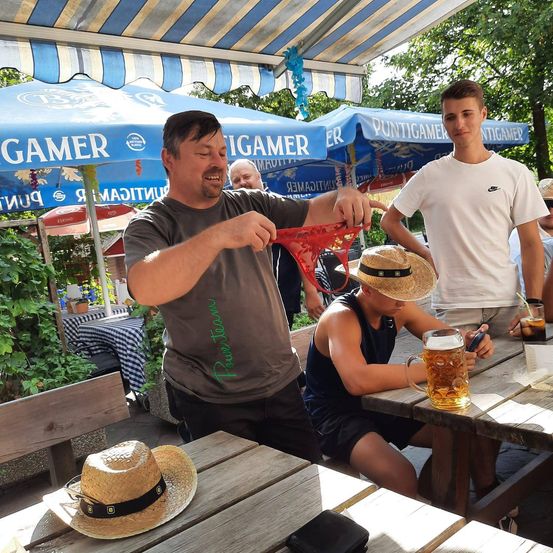 Ein Mann hält ein rotes Objekt und lächelt, mit anderen um ihn herum in einem Biergarten. Eine Frau in einem Hut sitzt in der Nähe, und ein Mann in einem weißen T-Shirt hält ein Handy.
