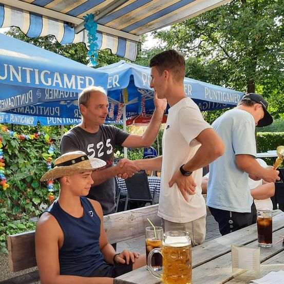 Drei Männer in einem Biergarten, einer schüttelt die Hand eines anderen, während ein Dritter ein Glas Bier in der Hand hält. Ein Junge in einem Strohhut sitzt auf einer Bank in der Nähe.