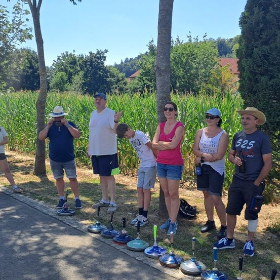 Eine Gruppe von Menschen steht in einem Maisfeld, trägt Hüte und Sonnenbrillen, und eine Person hält eine Bierflasche. Sie stehen neben einem Baum und einer Reihe von Rasenkegelpins.