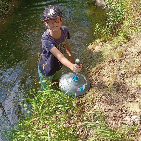 Ein junger Junge mit einer Mütze und einer Brille steht in einem Fluss und hält ein blaues und silbernes Objekt.