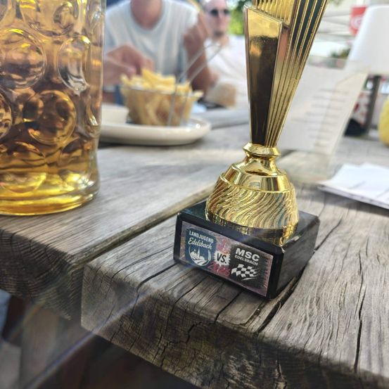Ein goldener Pokal mit schwarzer Basis steht auf einem Holztisch neben einem Bierglas. Im Hintergrund isst eine Person Pommes.