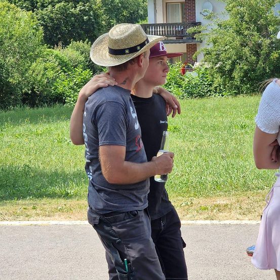 Zwei Männer und eine Frau gehen auf einem Bürgersteig. Der Mann in der Mitte hält eine Flasche. Der Mann auf der linken Seite trägt einen Strohhut.