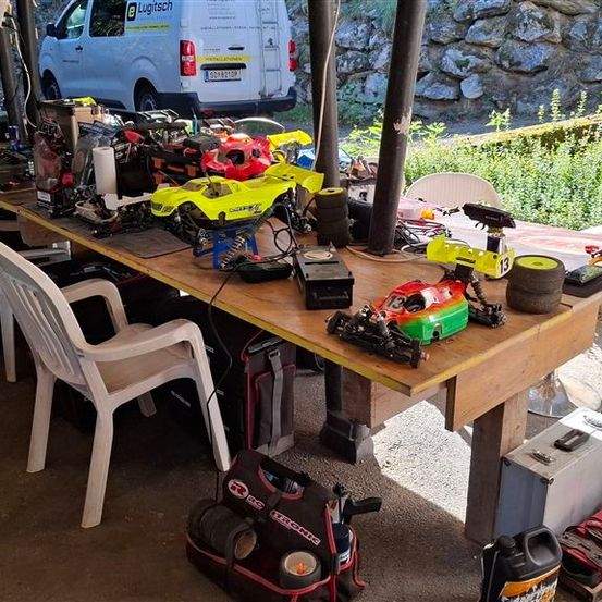 Ein hölzerner Tisch im Freien ist mit verschiedenen ferngesteuerten Autos, Werkzeugen und Ausrüstungsgegenständen übersät. Ein weißer Van ist dahinter geparkt und eine Steinmauer ist im Hintergrund.