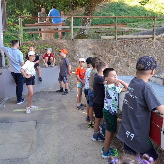 Eine Gruppe von Kindern und Erwachsenen steht auf einem Betonweg und beobachtet etwas vor sich. Die Kinder tragen Mützen und Turnschuhe. Hinter ihnen trennt ein Holzzaun sie von einem Grasbereich mit einem Baumstamm.