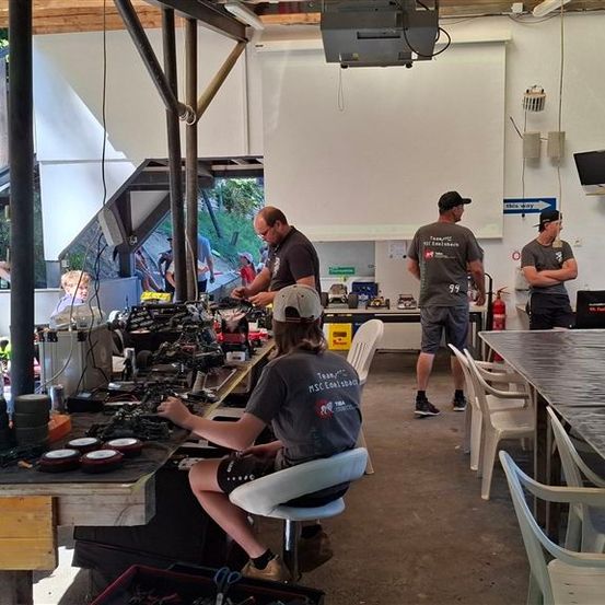 Eine Gruppe von Menschen arbeitet in einer Werkstatt an einem Projekt. Eine Frau sitzt auf einem Stuhl und arbeitet an einer Maschine, während andere stehen und zusehen.