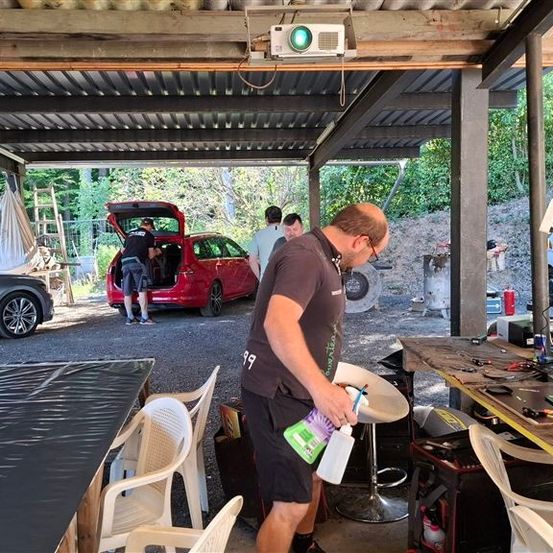 Ein Mann reinigt einen Tisch in einer Carport mit einer Sprühflasche, während zwei Männer Gegenstände in ein rotes Auto laden.
