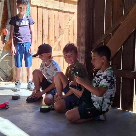 Vier Jungen spielen mit einem ferngesteuerten Auto in einer Scheune. Drei Jungen sitzen auf dem Boden und ein Junge steht.