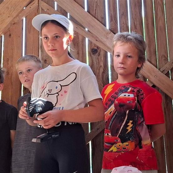Vier Kinder stehen vor einem Holzzaun. Das Mädchen in der Mitte hält einen Pokal. Der Junge rechts trägt ein rotes Lightning McQueen-Shirt.