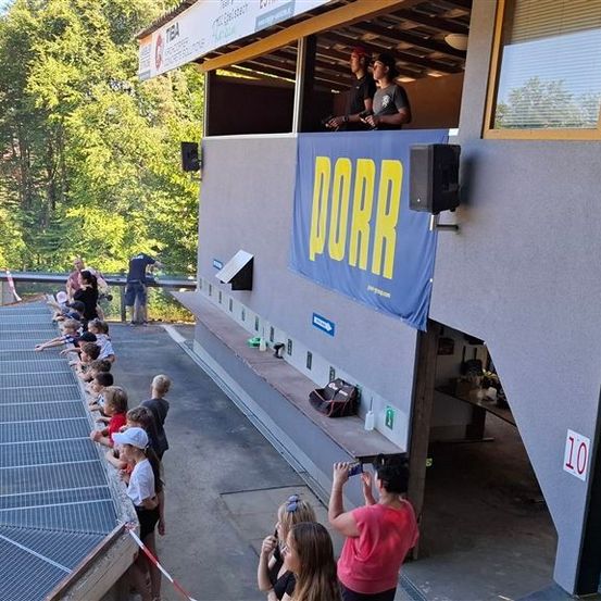 Eine Gruppe von Kindern und Erwachsenen steht in einer Schlange und schaut sich ein Sportereignis an. Das Event wird von Dorr gesponsert. Die Leute machen Fotos und Videos des Events.