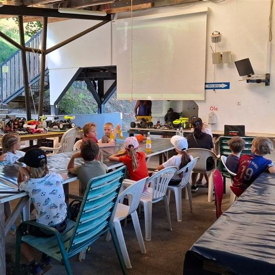Eine Gruppe von Kindern sitzt um einen Tisch in einem Raum mit einer Projektionsleinwand und Spielzeug auf dem Tisch.