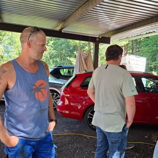 Zwei Männer stehen unter einem Carport, einer trägt ein ärmelloses Shirt mit Sonnen- und Palmendesign, der andere ein helles Shirt. Ein rotes Auto ist in der Nähe geparkt, und eine Frau steht rechts.