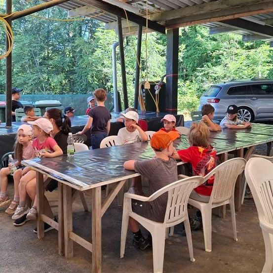 Eine Gruppe von Kindern und Erwachsenen sitzt an einem langen Tisch unter einem Schutzdach, mit Bäumen und einem Auto im Hintergrund.