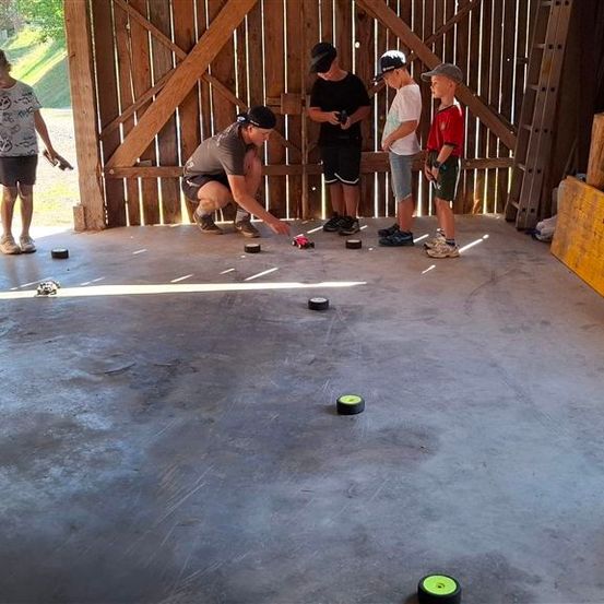 Eine Gruppe von Kindern und einem Erwachsenen spielen ein Spiel mit kleinen Autos auf einem Betonboden. Der Erwachsene hockt sich hin und scheint den Kindern etwas zu erklären.