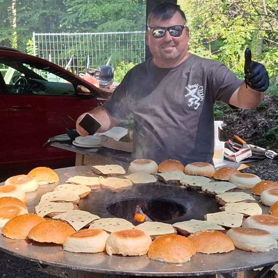 Ein Mann mit Sonnenbrille und Handschuhen steht neben einem großen Grill mit mehreren Sandwiches darauf. Er hält ein Handy und gibt einen Daumen hoch.