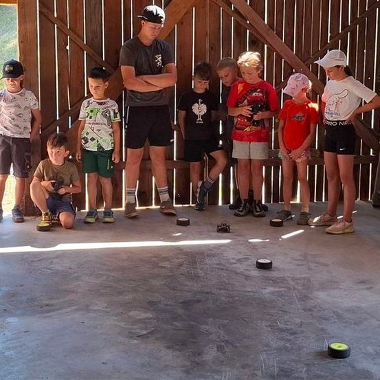 Eine Gruppe von Kindern und einem Mann stehen in einer Garage mit einer Holzwand, einige knien und andere stehen mit verschränkten Armen. Sie spielen ein Spiel mit kleinen Objekten auf dem Boden.