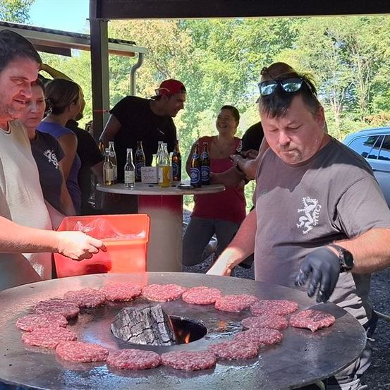 Eine Gruppe von Menschen versammelt sich um einen großen Grill im Freien und kocht Hamburger. Ein Mann zeigt auf die Patties, während ein anderer Mann mit Sonnenbrille grillt.