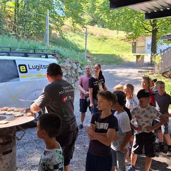 Eine Gruppe von Kindern und Erwachsenen beobachtet einen Mann, der Essen auf einem Grill zubereitet. Der Mann trägt ein graues Shirt mit Logo. Die Kinder tragen Mützen und Turnschuhe. Der Mann hält einen Teller mit Essen.