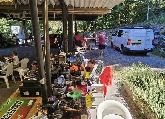 Eine Gruppe von Menschen versammelt sich um einen Tisch mit verschiedenen Werkzeugen und Ausrüstungsgegenständen, möglicherweise zur Vorbereitung eines Autorennens. Ein weißer Lieferwagen ist in der Nähe geparkt, und mehrere Autos sind am Straßenrand geparkt.