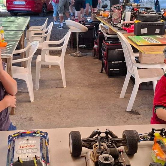 Zwei junge Jungen spielen mit ferngesteuerten Autos bei einer Outdoor-Veranstaltung. Ein Junge trägt einen Hut und der andere trägt ein Shirt mit einem Logo. Im Hintergrund befinden sich Stühle, Tische und ein Auto.